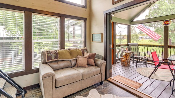 A cozy living area with a leather sofa, a wooden coffee table, and a red chair. There is a large window with blinds, a door leading to an outdoor deck, and a welcome mat on the floor. - 1 Mi to Tank Town Usa: Tiny Home w/ Pool Access! (Morganton)