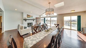 A spacious living room with a fireplace, a dining area with a long table, and a view through large windows. - Waterfront Home w/ Dock, Hot Tub, Fire Pit (Oak Harbor)