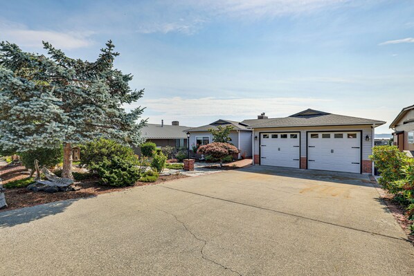A residential driveway leading to a house with a garage, surrounded by landscaped greenery and a large tree.