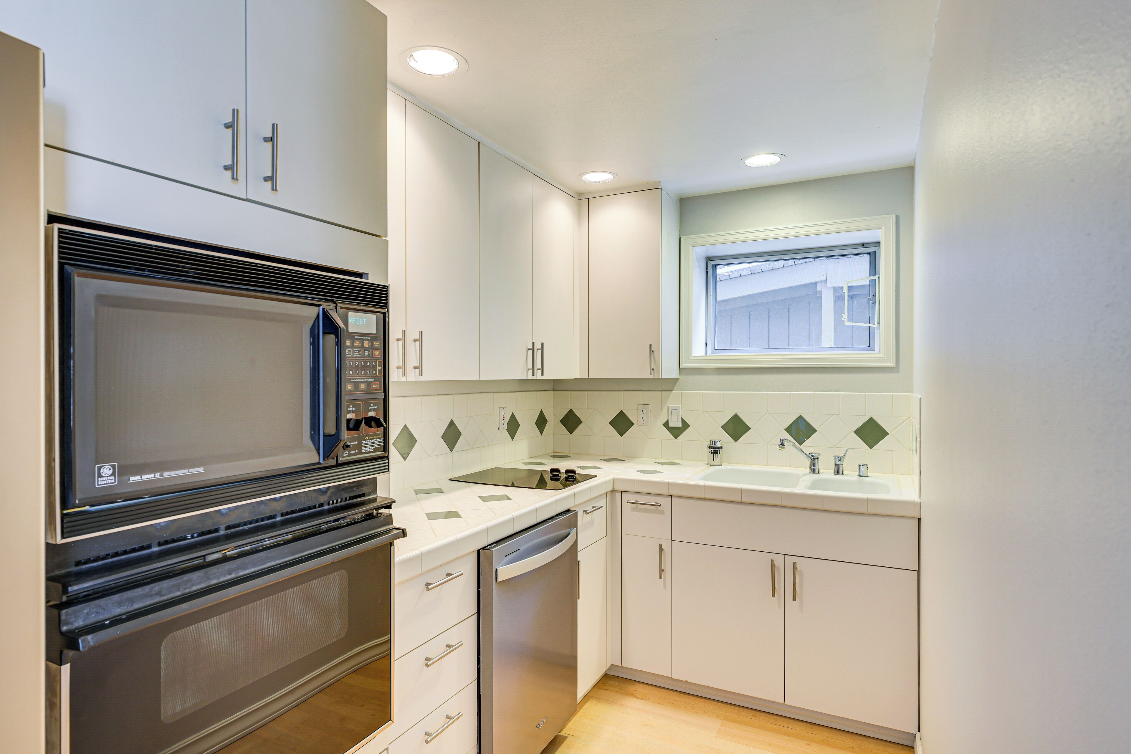 A modern kitchen with white cabinets, a built-in microwave, a dishwasher, and a window above the sink.