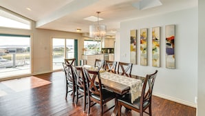 A spacious dining area with a large table, multiple chairs, and a chandelier. There are large windows offering a view of the outdoors. - Waterfront Home w/ Dock, Hot Tub, Fire Pit (Oak Harbor)