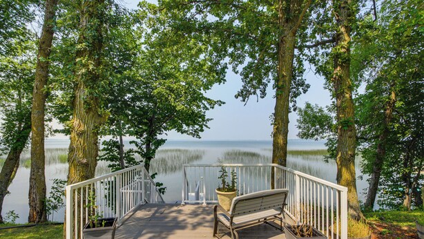 Une terrasse en bois avec un banc et des plantes en pot, donnant sur un lac paisible bordé d’arbres.