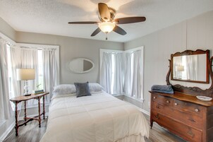 Un dormitorio con cama, cómoda, espejo, ventilador de techo y ventana con cortinas.