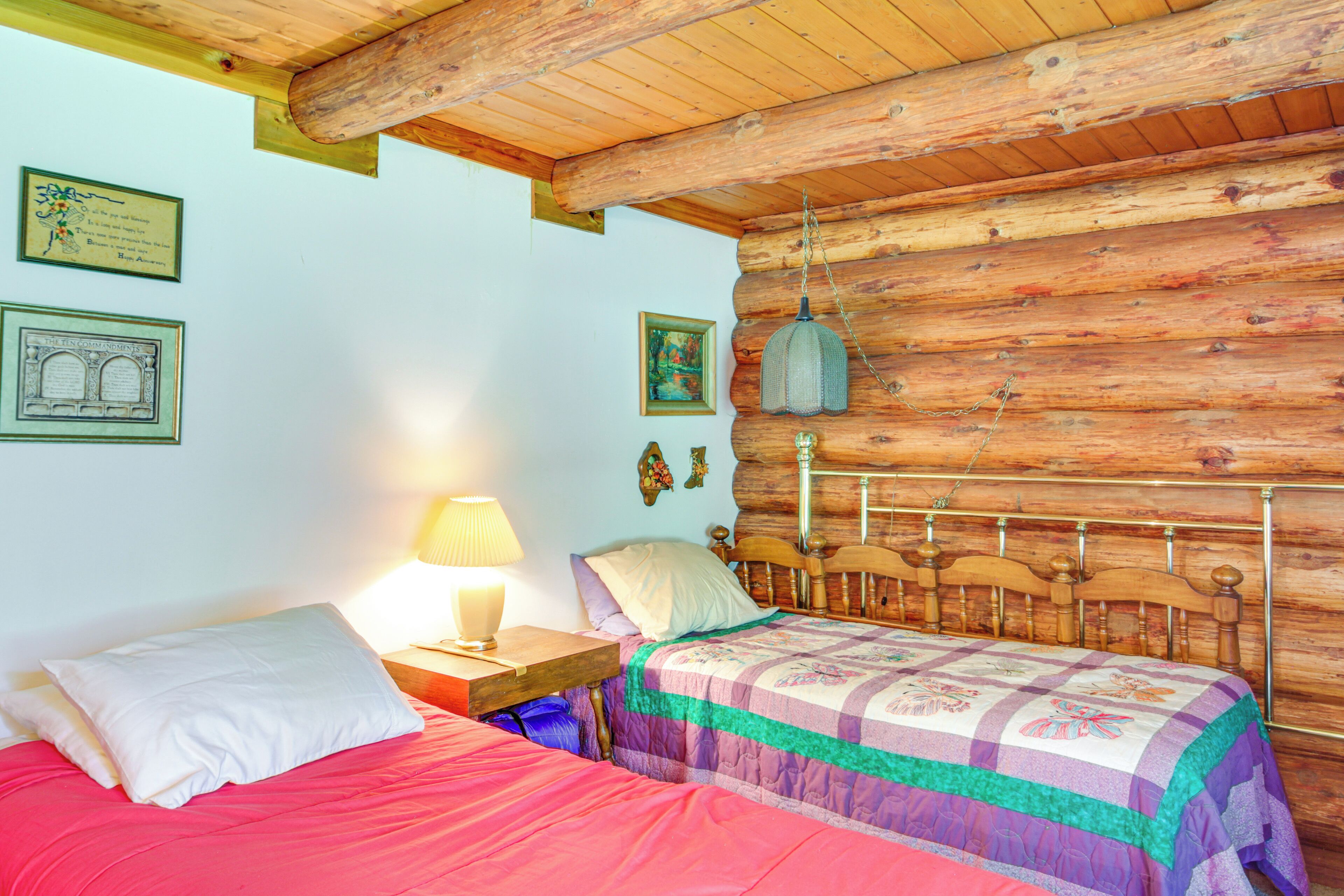 Een gezellige kamer met een houten wand, een bed met een kleurrijke quilt, een nachtkastje met een lamp, en ingelijste foto’s aan de muur.