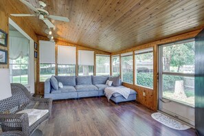 Una habitación acogedora con techo de madera, un sofá modular gris, un sillón de mimbre y amplias ventanas con vistas al exterior.