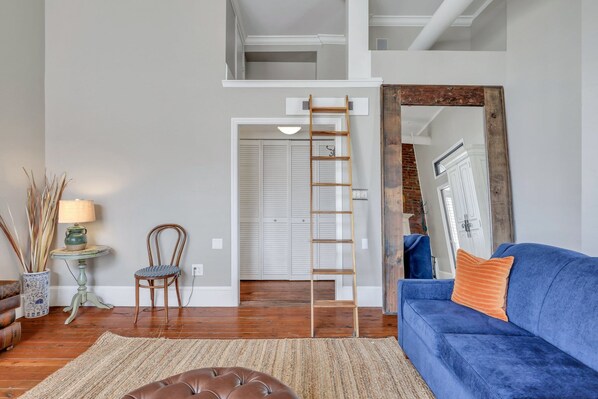 Una sala de estar con un sofá azul, un reposapiés marrón, una escalera de madera, un espejo y una puerta que da a un armario.