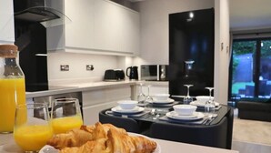 A modern kitchen with a dining area, breakfast setup with croissants and orange juice, and a view of the outdoors. - Spotless Two-Bedroom Home with Garden and Parking (Hertfordshire)