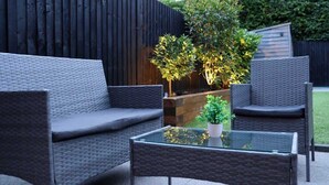 An outdoor seating area with wicker furniture and a glass table, surrounded by a wooden fence and greenery. - Spotless Two-Bedroom Home with Garden and Parking (Hertfordshire)