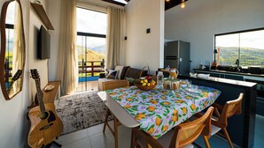 A spacious living area with a dining table, chairs, and a guitar. A kitchen is visible in the background. - Chalé Arca da Vida (Piquete)