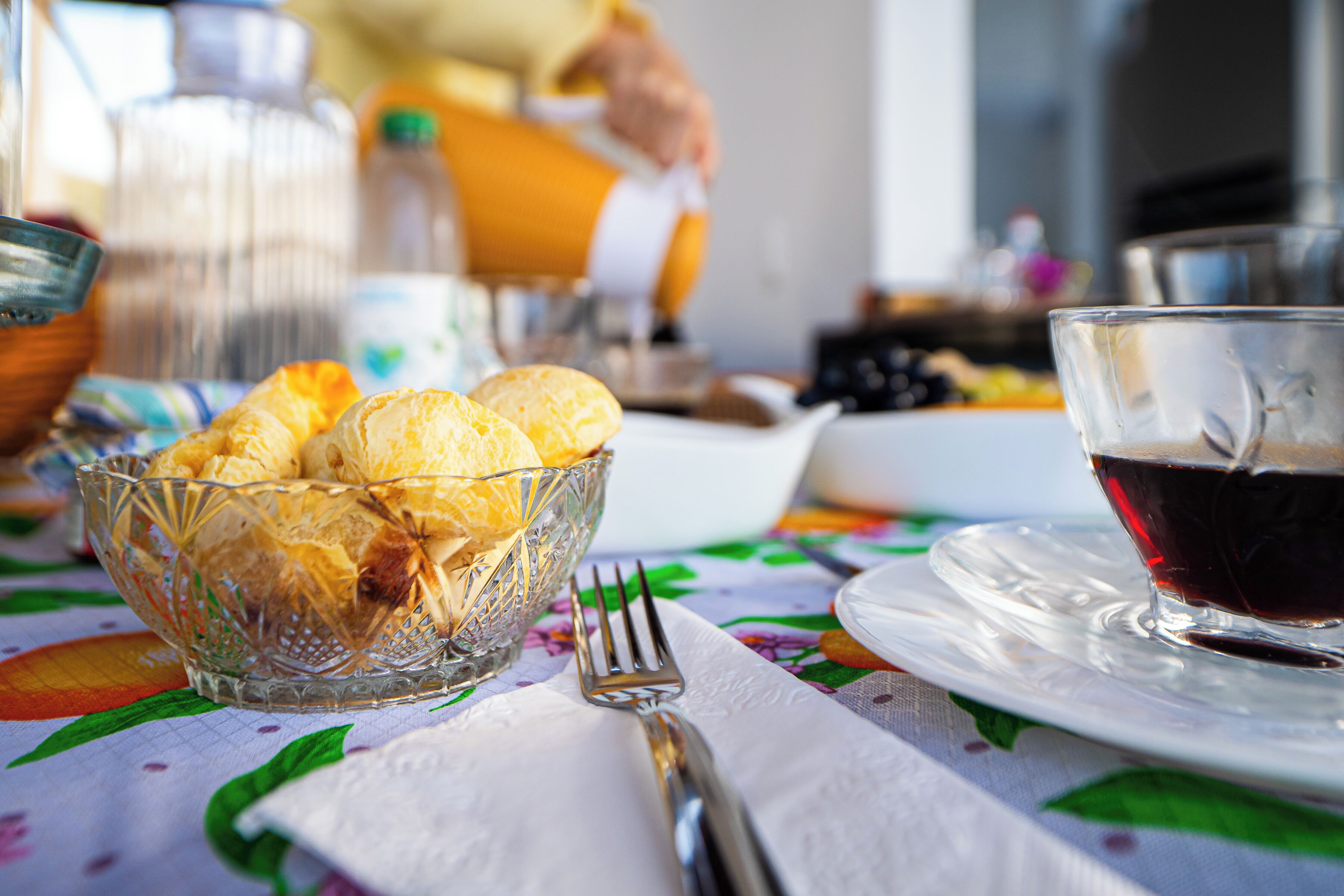 Une table mise pour le petit-déjeuner, avec un saladier en verre rempli de viennoiseries, une tasse de café et une fourchette, le tout posé sur une table recouverte d’une nappe à motifs floraux.