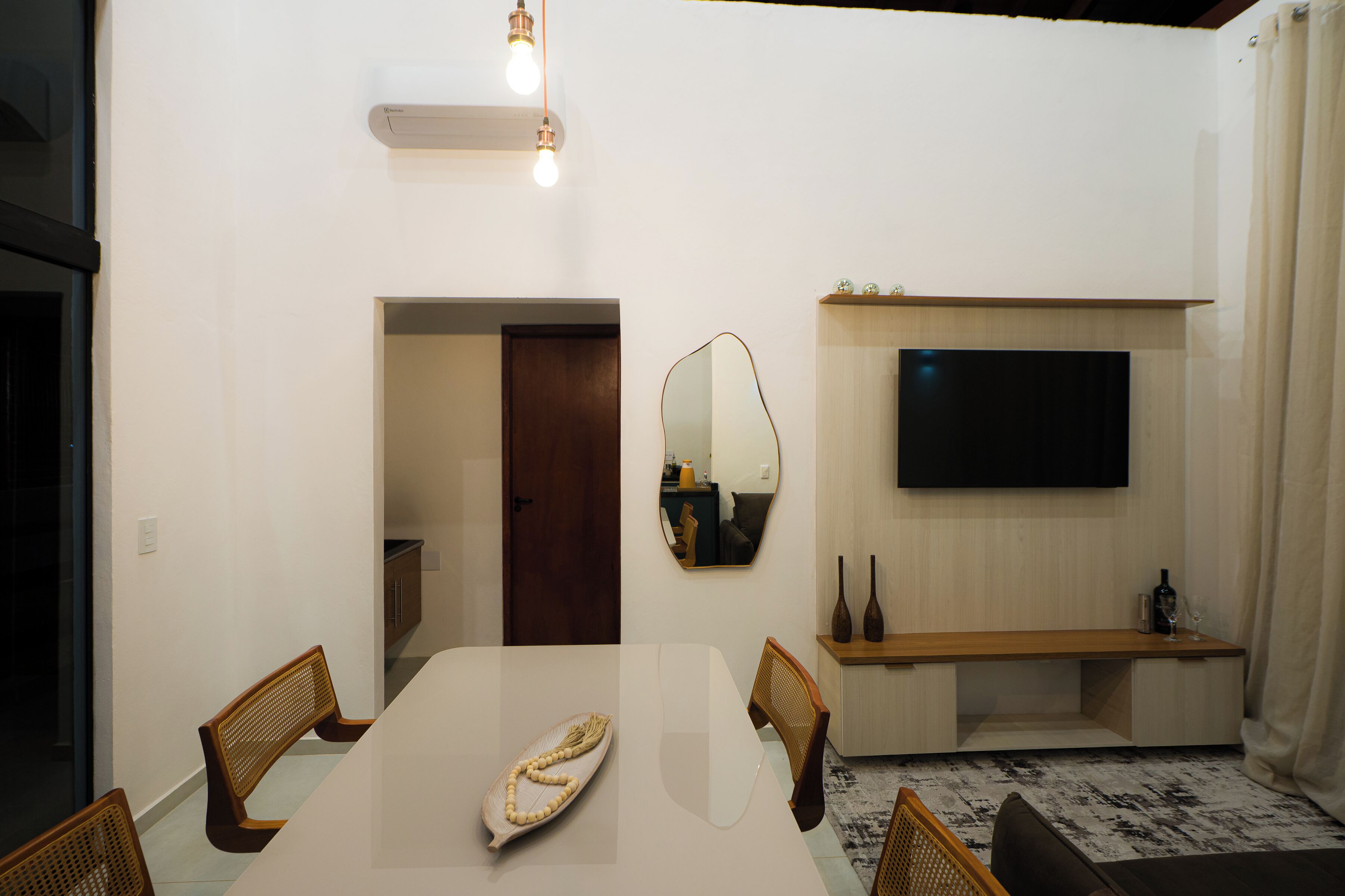 Un salón moderno con una mesa de comedor blanca, dos sillas de mimbre, un televisor de pantalla plana en la pared y un banco de madera con una copa de vino y dos botellas.