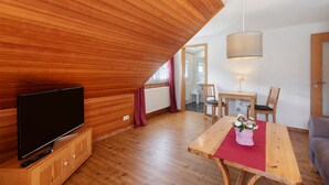A room with a TV on a wooden cabinet, a dining table with chairs, a hanging light fixture, and a wooden ceiling.