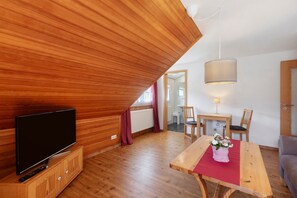A room with a TV on a wooden cabinet, a dining table with chairs, a hanging light fixture, and a wooden ceiling.