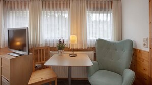 Une pièce avec un bureau en bois, une télévision, une lampe, une chaise et une fenêtre avec des rideaux.