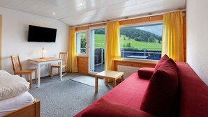 Quarto com cama, sofá vermelho, mesa de madeira, televisão na parede e janela com vista para uma paisagem verde.