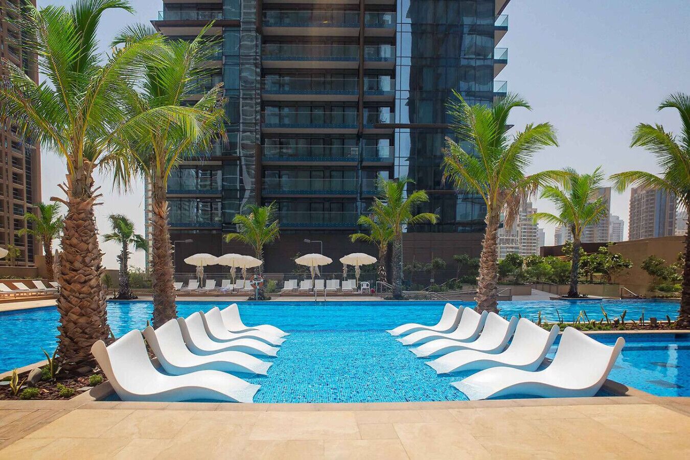 A swimming pool with white lounge chairs and palm trees, surrounded by tall buildings.