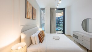 A modern hotel room with a large bed, a bedside lamp, a dresser, and a view of the pool and buildings outside. - Binghatti Crescent - Two Bed with Pool View in JVC (Jumeirah Village Circle)