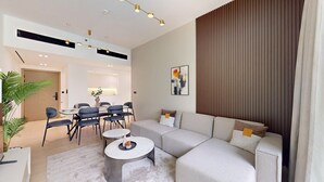A modern living room with a sectional sofa, a dining area with a table and chairs, and a large window with blinds. - Binghatti Crescent - Two Bed with Pool View in JVC (Jumeirah Village Circle)