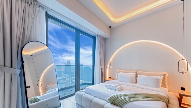 Quarto moderno com uma cama grande, um espelho curvo e vista da cidade através de janelas do chão ao teto.