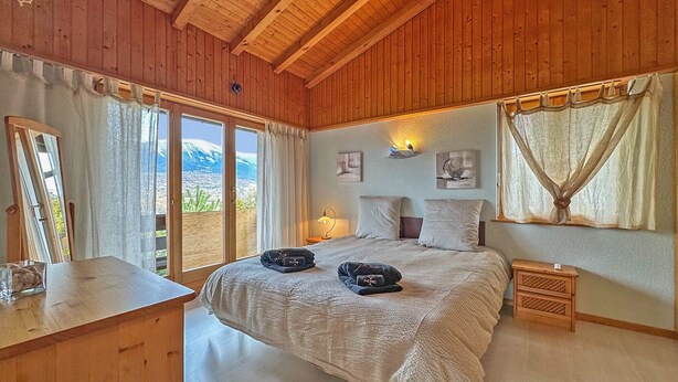 Ein Schlafzimmer mit Holzboden, einem Bett mit weißer Decke, einem Nachttisch, einem Spiegel und Blick auf Berge durch das Fenster.