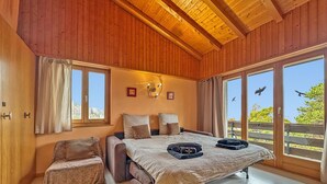 Ein gemütliches Schlafzimmer mit Bett, einer kleinen Sitzecke und großen Fenstern mit Blick ins Freie.