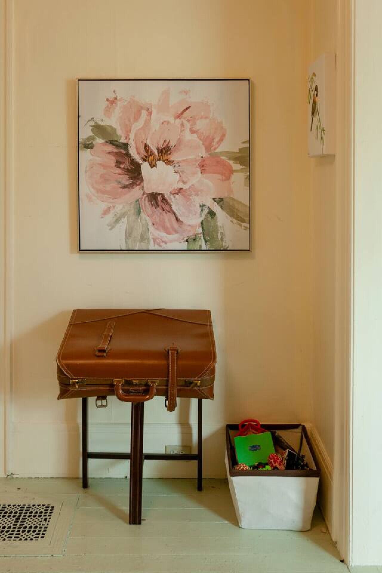 A room with a brown leather chair, a white storage bin, and a framed floral painting on the wall.