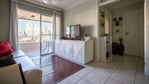 A modern living room with a sofa, a TV on a white cabinet, a city view through large windows, and a ceiling fan.
