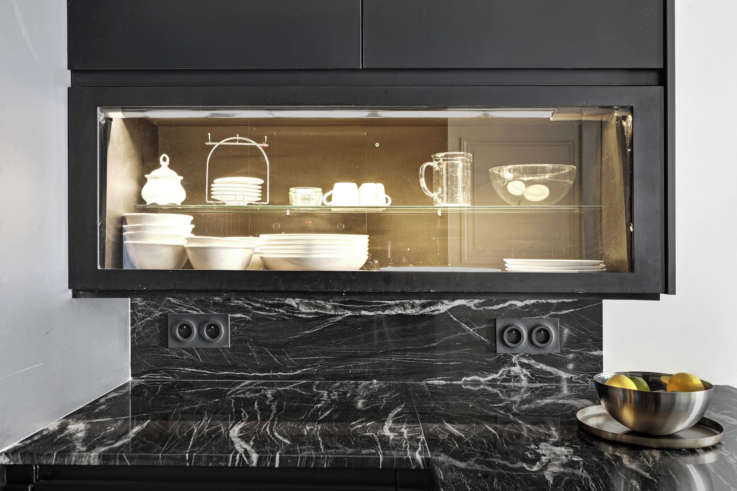 A kitchen with a black marble countertop, a glass display cabinet containing white dishes, and a bowl of lemons on the counter.