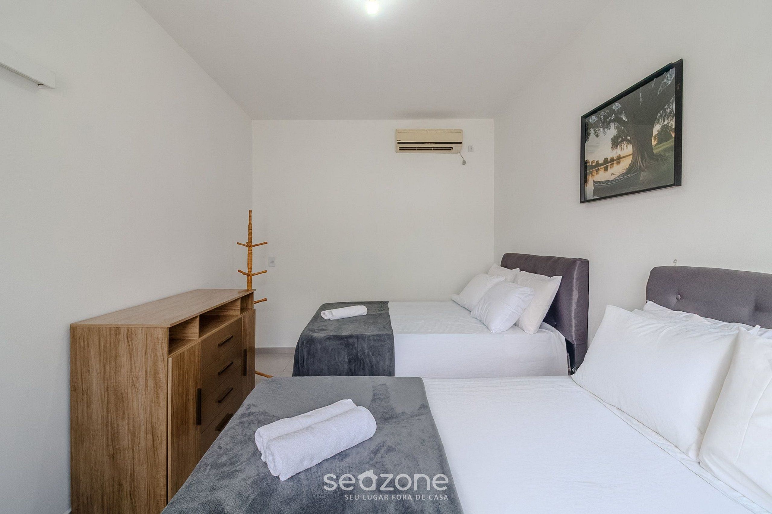 Habitación de hotel con dos camas, un aparador de madera, una mesita con una toalla, aire acondicionado y un cuadro en la pared.