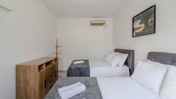 Habitación de hotel con dos camas, un aparador de madera, una mesita con una toalla, aire acondicionado y un cuadro en la pared.