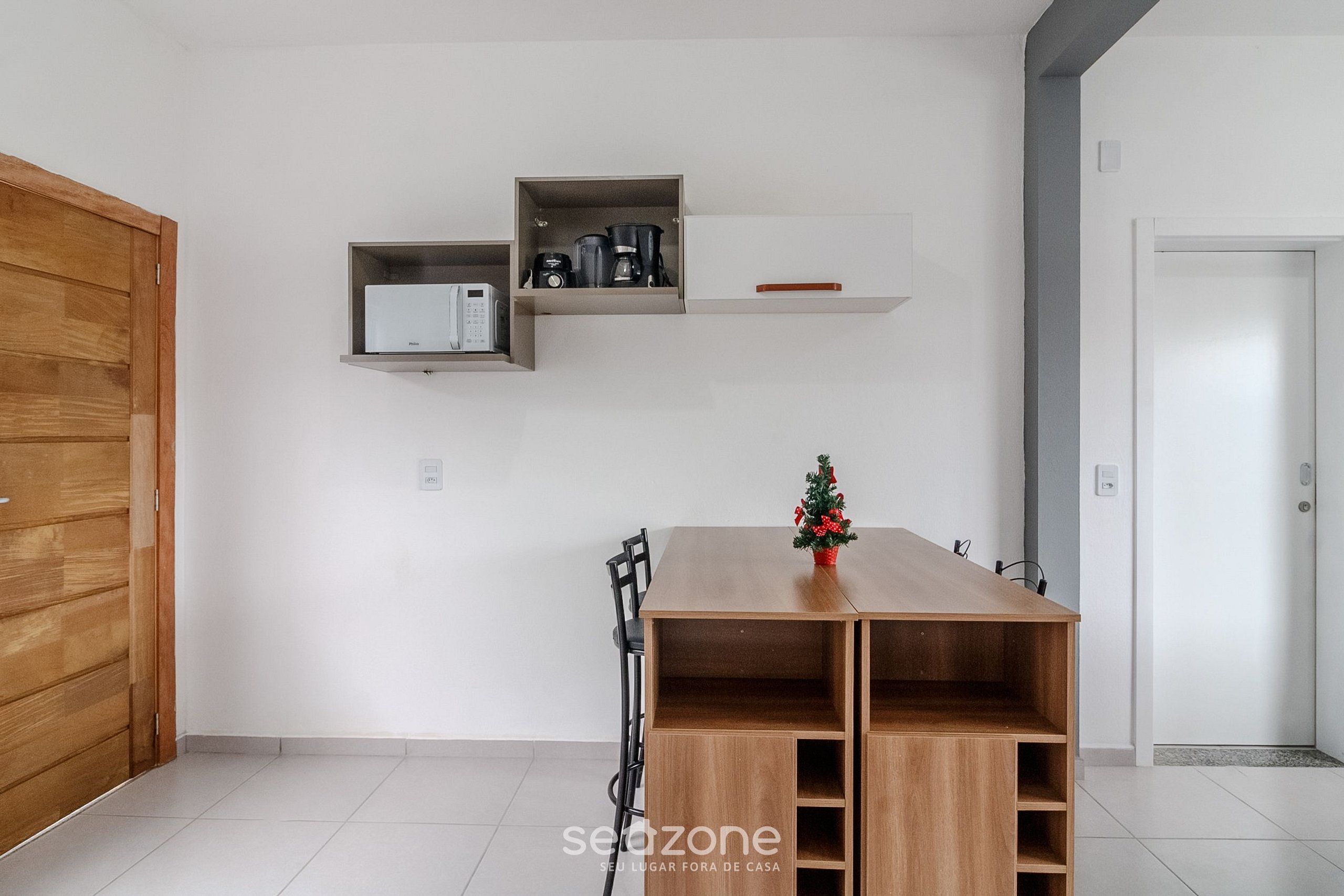Una habitación moderna con una mesa de madera, un pequeño árbol de Navidad y estantes empotrados con electrodomésticos de cocina.