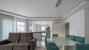 Una sala de estar moderna con un sofá modular, una mesa de comedor de vidrio y sillas color turquesa. Se aprecia una cocina al fondo, con armarios blancos y un refrigerador de acero inoxidable.