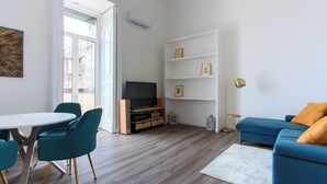 A modern living room with a TV, blue sofa, teal chairs, and a white wall with shelves.