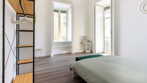 A bedroom with a bed, a wooden shelf unit, a window, and a door leading to another room.