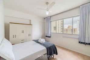Quarto com cama, guarda-roupa, janela com cortinas e ventoinha no teto.