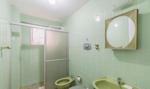 Casa de banho com azulejos verdes, cabine de duche envidraçada, duas sanitas, uma pia e um espelho.