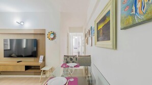 Una sala de estar moderna con una televisión de pantalla plana, un comedor con una mesa dispuesta para dos personas, y un pasillo con obras de arte enmarcadas.