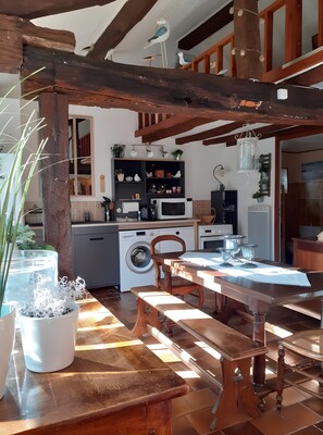 A rustic kitchen with wooden beams, a washing machine, and a dining area with a wooden table and benches.