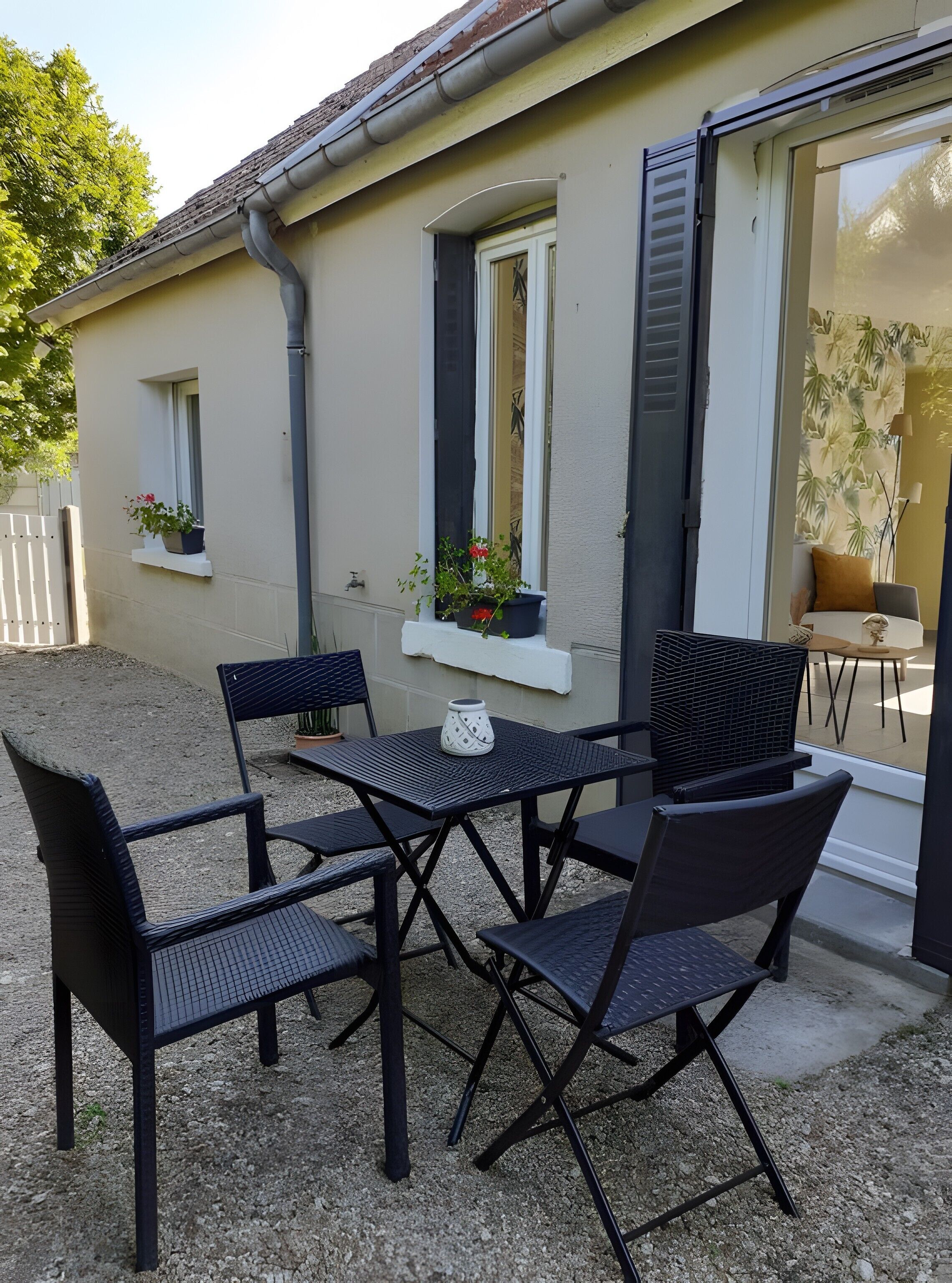 An outdoor seating area with a table and chairs, a potted plant, and a window with shutters.