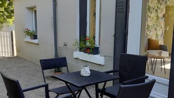 An outdoor seating area with a table and chairs, a potted plant, and a window with shutters.
