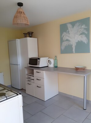 A kitchenette with a microwave, kettle, and a bowl on the counter, a refrigerator, a cabinet with drawers, and a hanging light fixture.