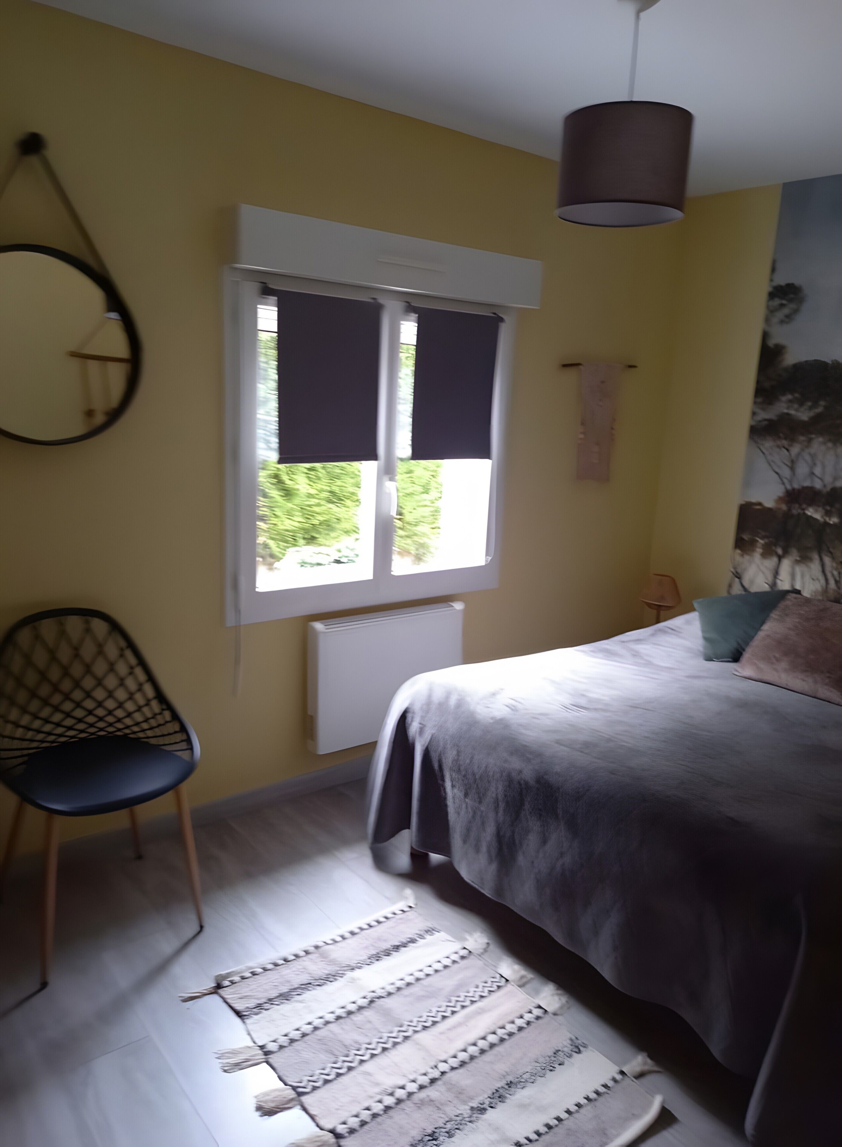 A bedroom with a bed, a chair, a window with blinds, a radiator, and a hanging light fixture.