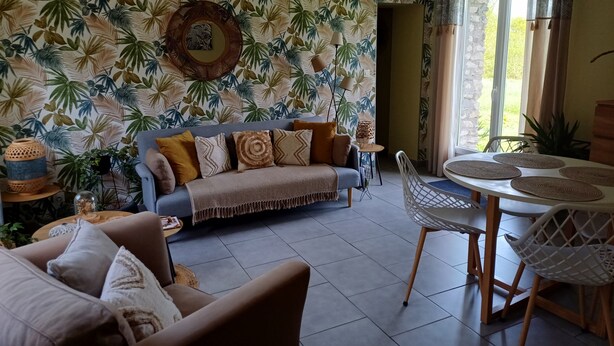 A living room with a tropical leaf-pattern wallpaper, a sofa with multiple cushions, a dining table with chairs, and a hanging light fixture.