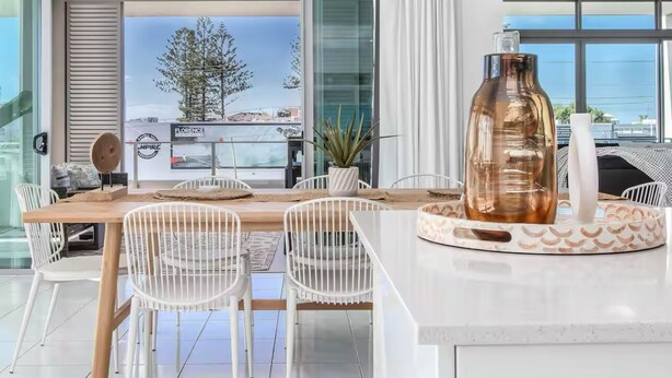 A modern dining area with a wooden table and white chairs, a large glass vase, and a view of the sea through large windows.