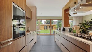 Eine moderne Küche mit Holzschränken, einem eingebauten Ofen und Blick auf eine grüne Wiese und ein Haus durch Glastüren.