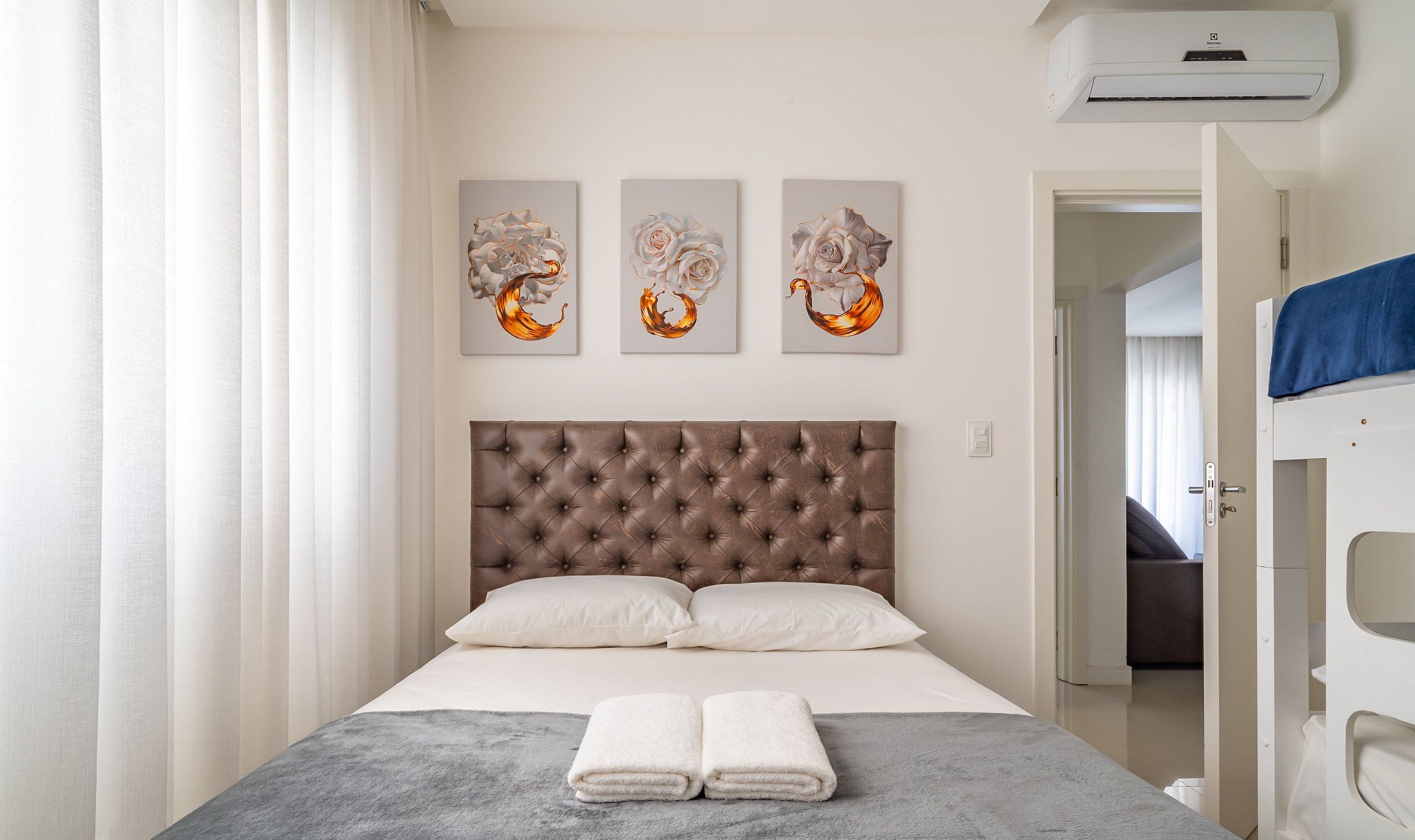 A neatly arranged hotel room with a tufted headboard, a bed with white linens, and three abstract paintings on the wall.