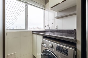 A compact kitchen with a built-in washer and dryer, a window with blinds, and a view of the cityscape. - Beautiful Apartment in Itajai Ctp1103 (Itajai)