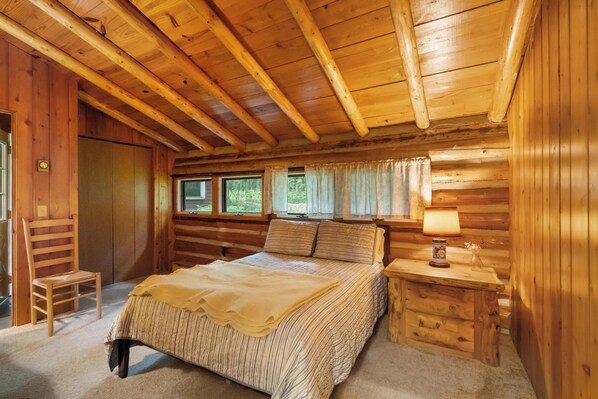 Une chambre dans un chalet en bois, avec un lit, une table de chevet, une chaise et une fenêtre avec des rideaux.