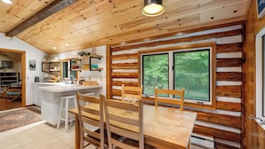 Een knus blokhutinterieur met een houten eettafel, stoelen, een keuken met witte kasten en een groot raam met uitzicht op het bos.