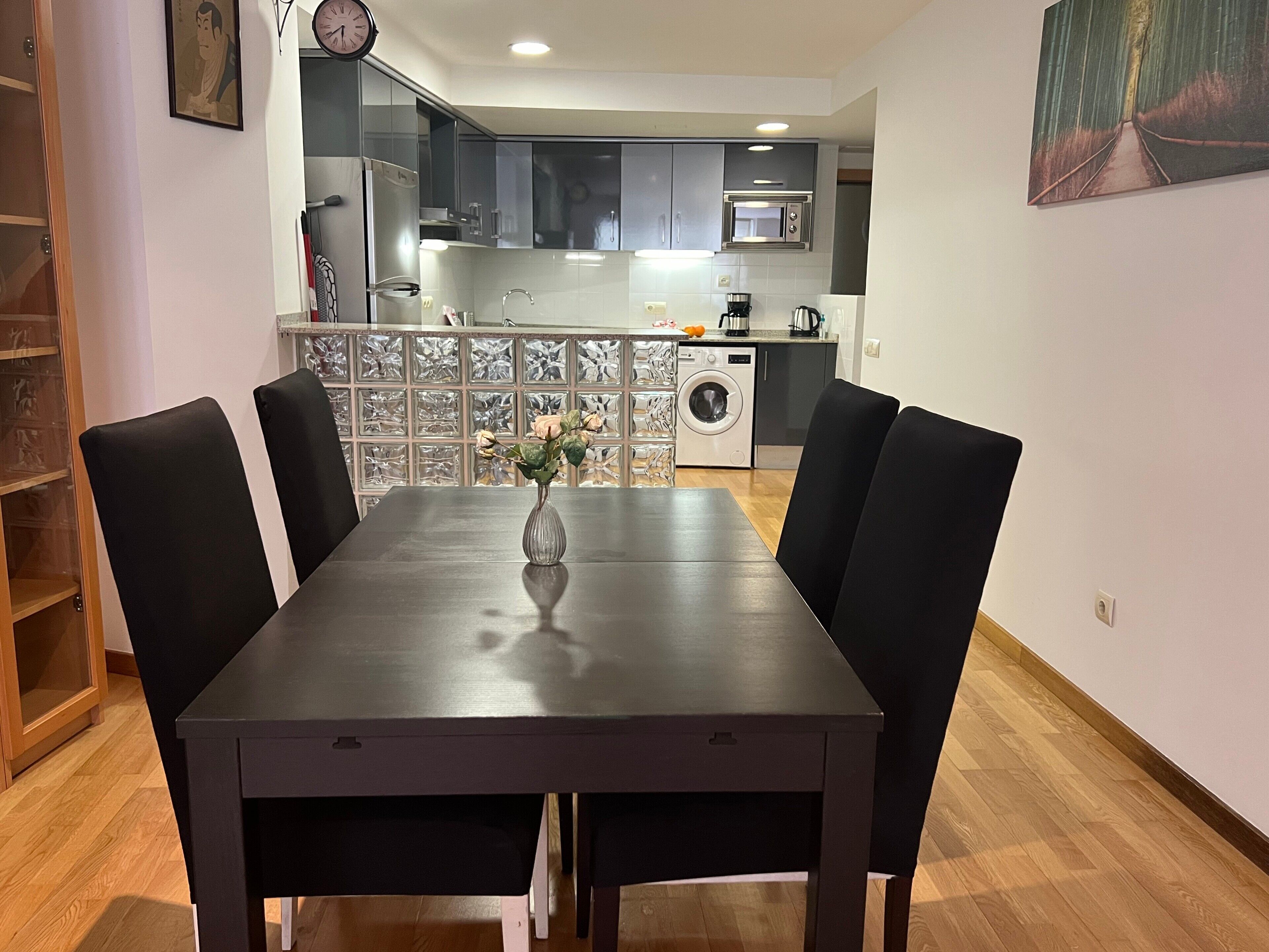 Una mesa de comedor con sillas, un florero con flores, un reloj y una cocina al fondo.